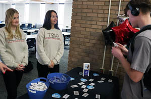 Samantha Ohnoutka and Jade Inlow visit with a student at a table in the cafeteria during the Student Support Services celebration of National Trio Day.