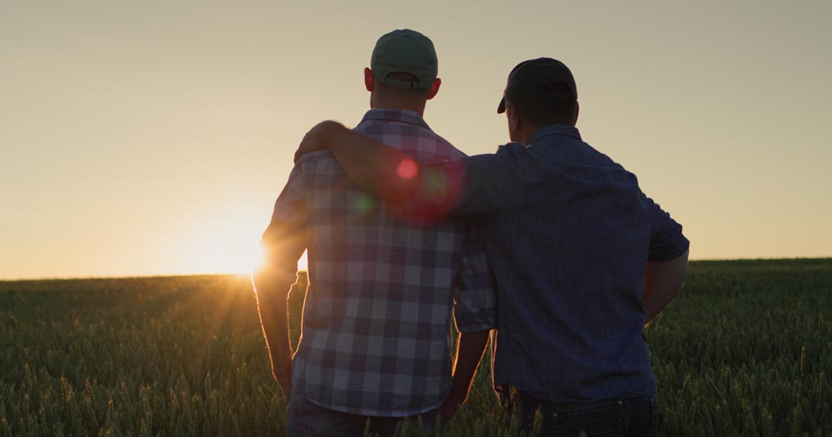 Farmer has his arm around his adult son in a wheat field as they watch a sunset.